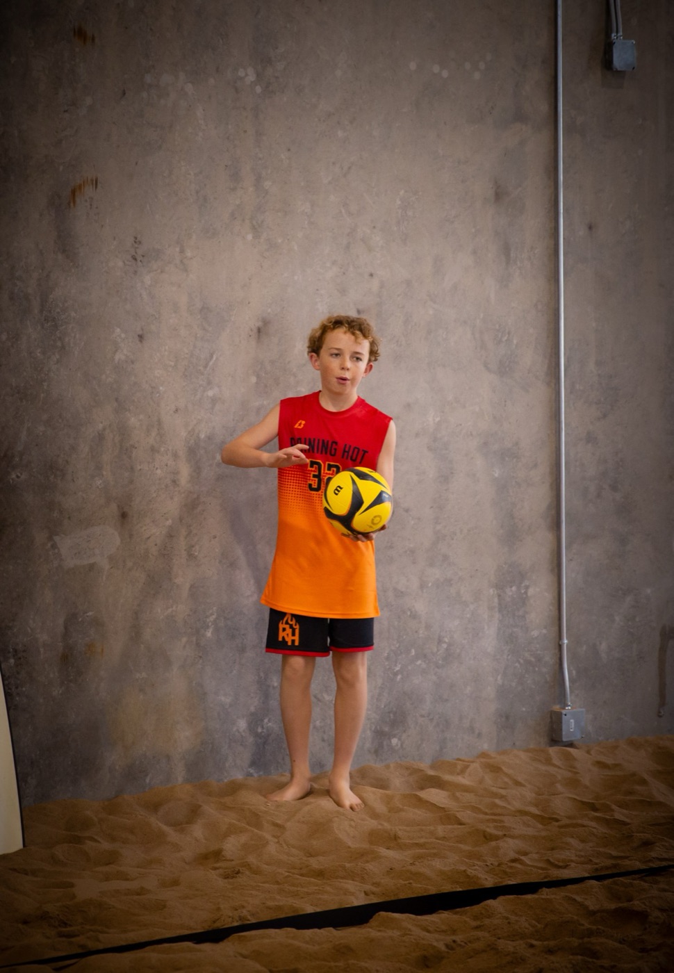 Young player in a red and orange jersey holding a volleyball on the sand